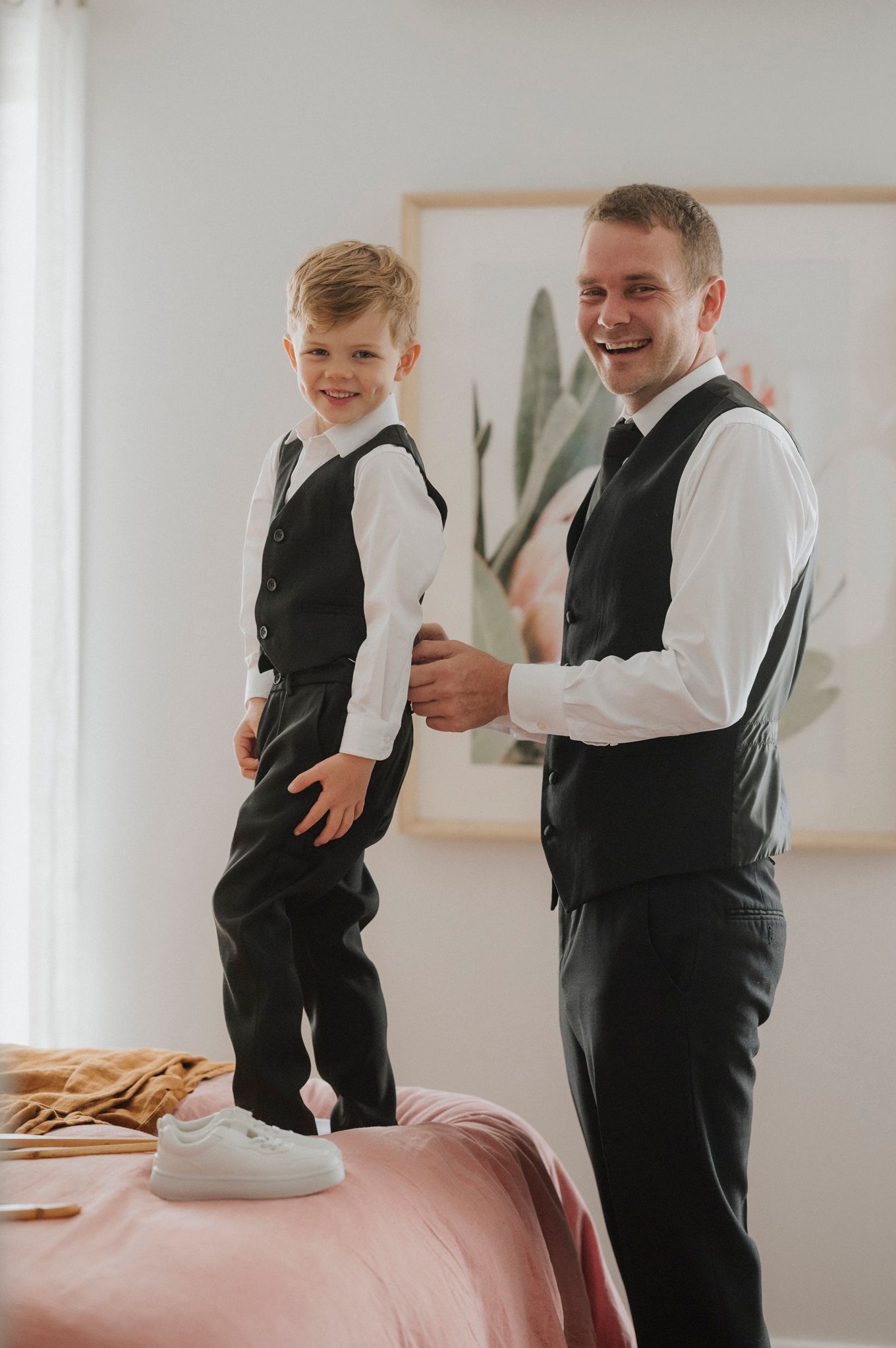 A man and young boy in matching black vests and white shirts smile together in a bright bedroom.