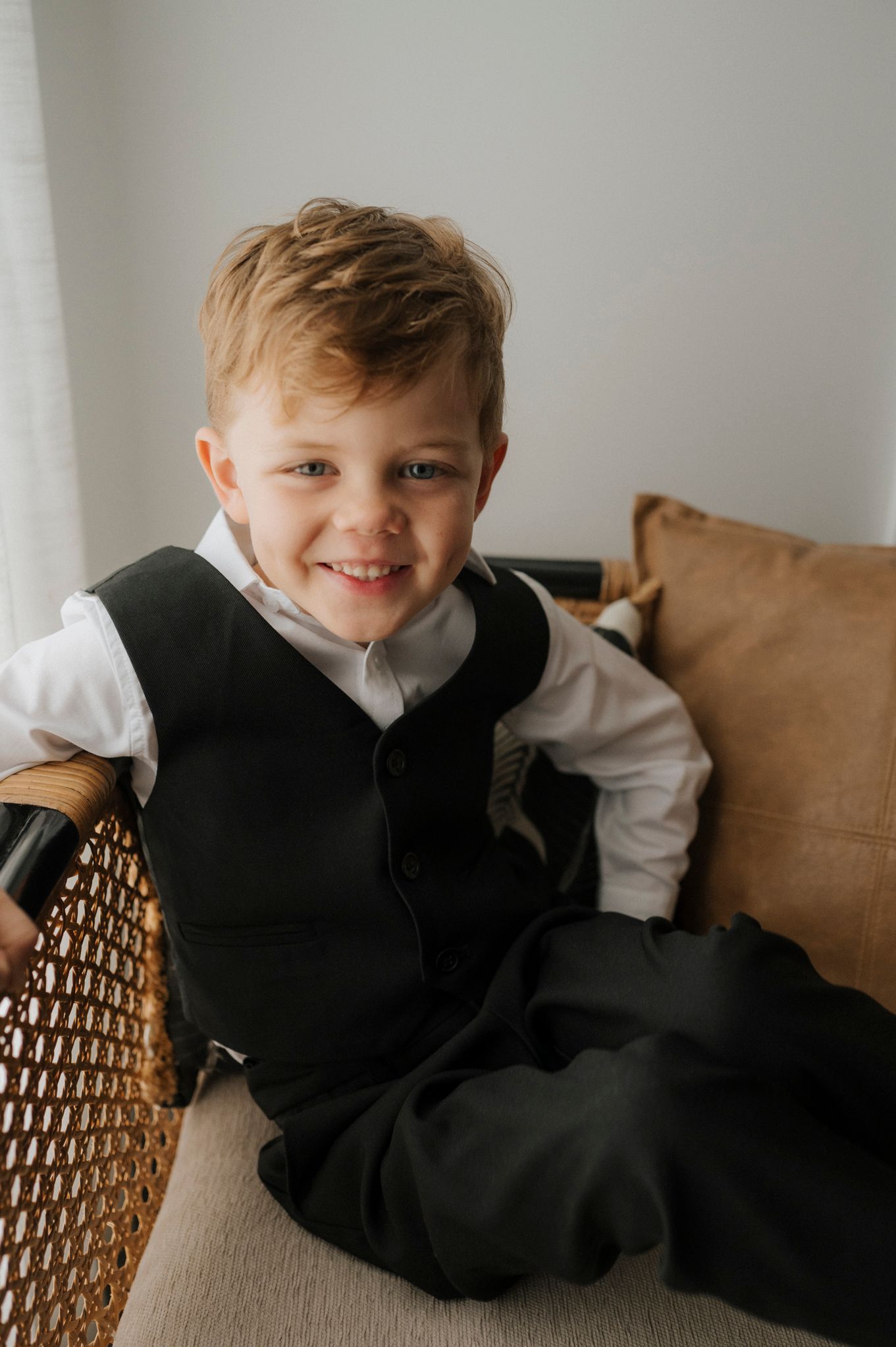 Smiling young boy in a black vest and white dress shirt sitting in a wicker chair at a formal event.