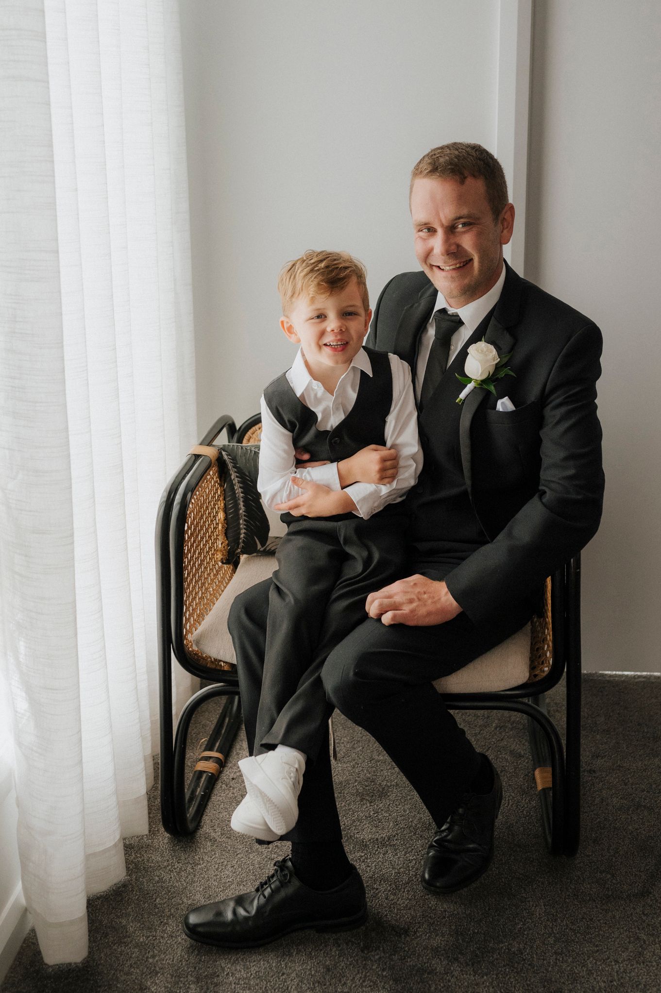 A groom in a black suit holds a young boy in matching attire, both smiling while seated by a window.