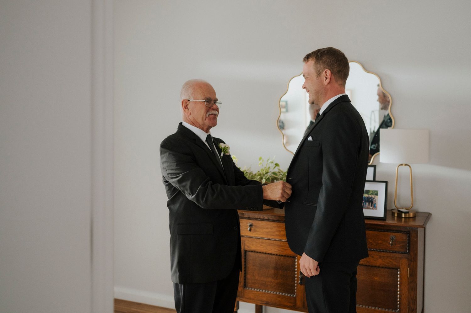 Two men in black suits share a moment as an older gentleman adjusts the younger mans boutonniere before a wedding.