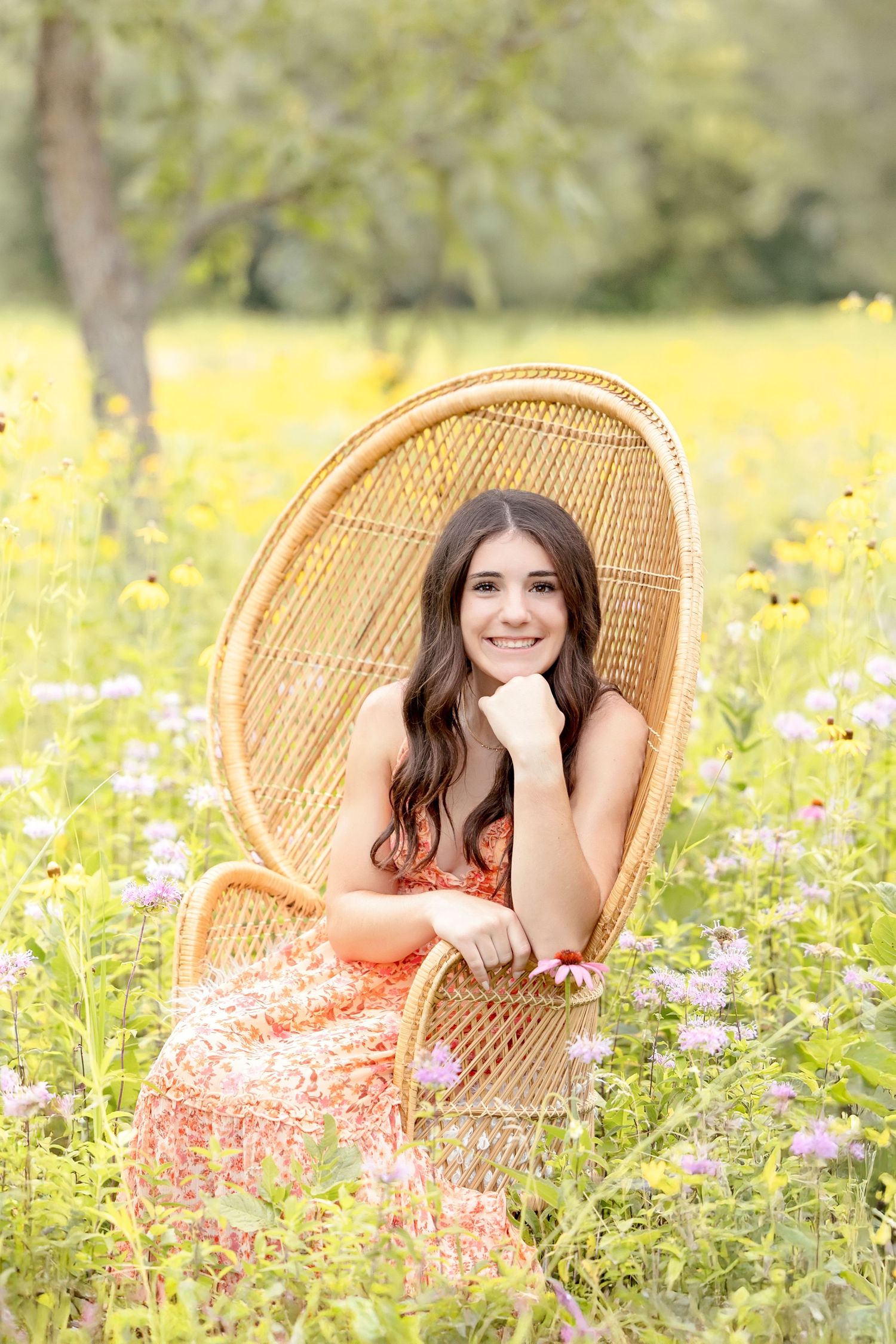 Senior girl sitting in a wicker chair surrounded by summer wildflowers during an outdoor senior photography session in North Iowa.