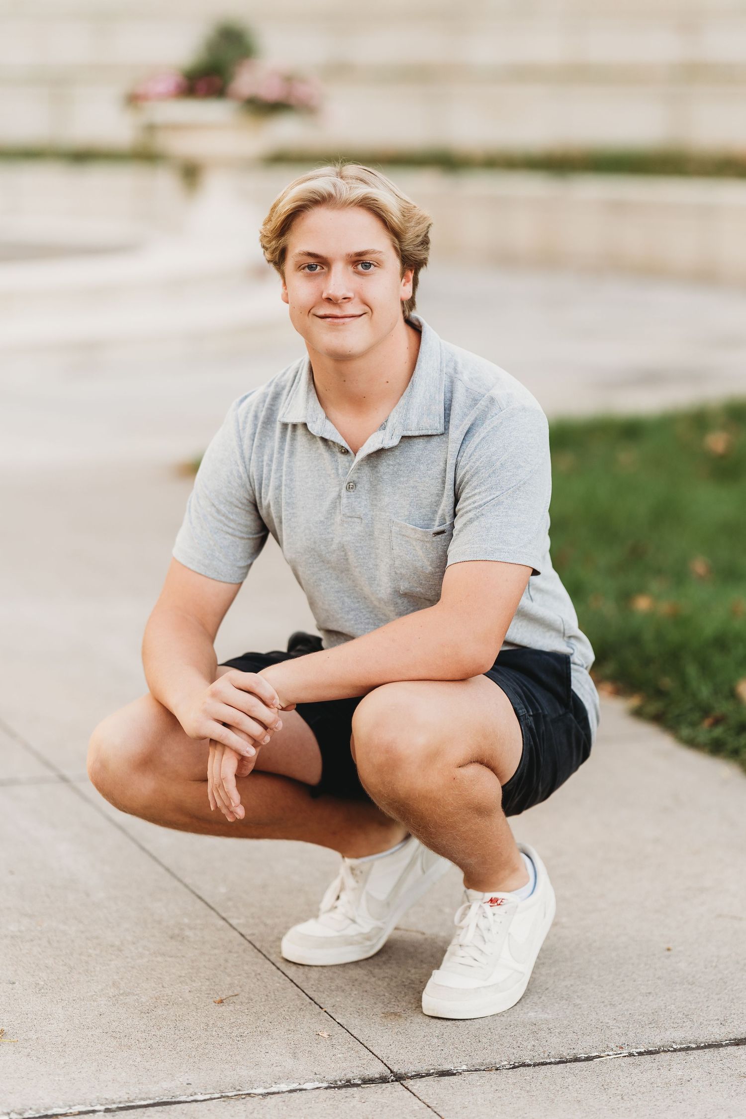 Young man in gray polo and dark shorts crouching on a sidewalk outdoors, smiling at the camera.