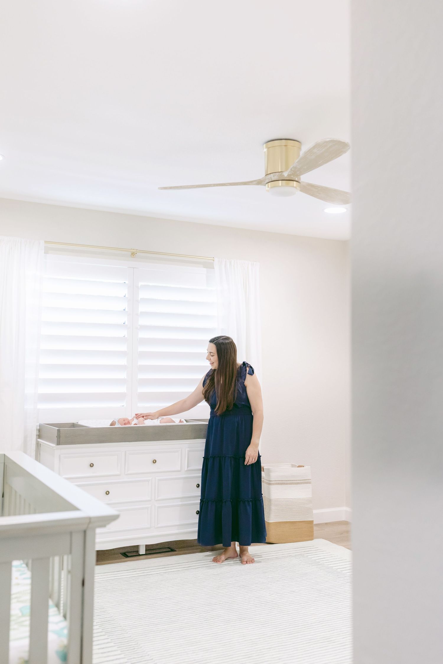 Woman in navy dress arranging items on a white dresser in a bright, modern nursery with ceiling fan.