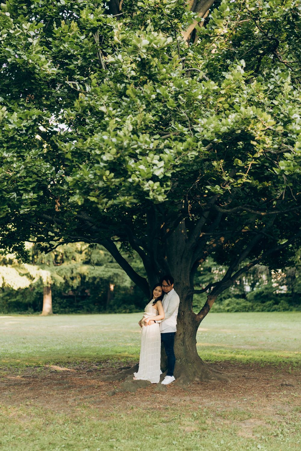 Expectant couple standing beneath a large tree in a green park.
