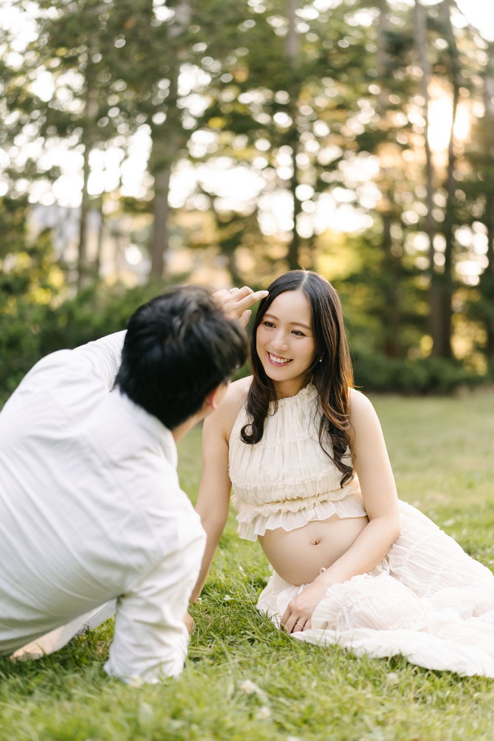 Partner kissing the expectant mother's cheek as they sit on the grass at sunset.
