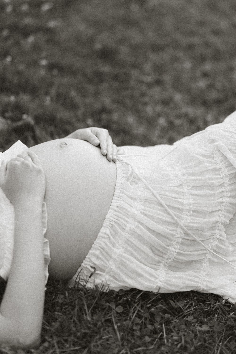 Close black-and-white detail of an expectant mother lying in a pleated dress on the grass.
