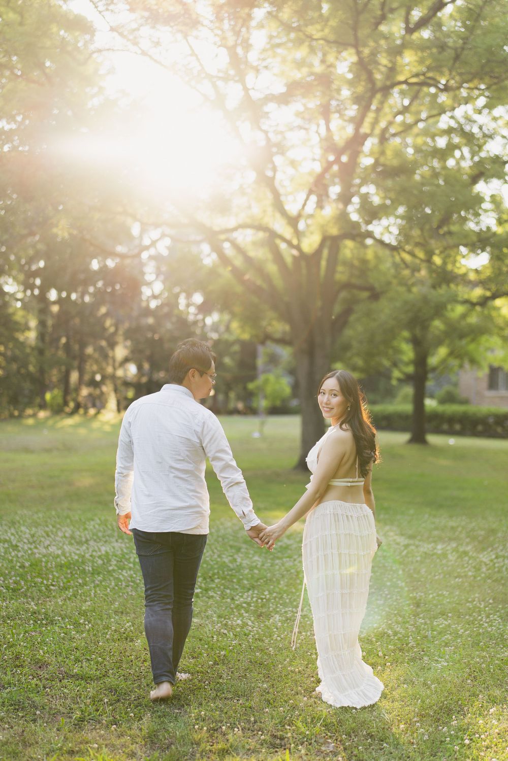 Expectant couple walking hand in hand across a sunlit lawn.