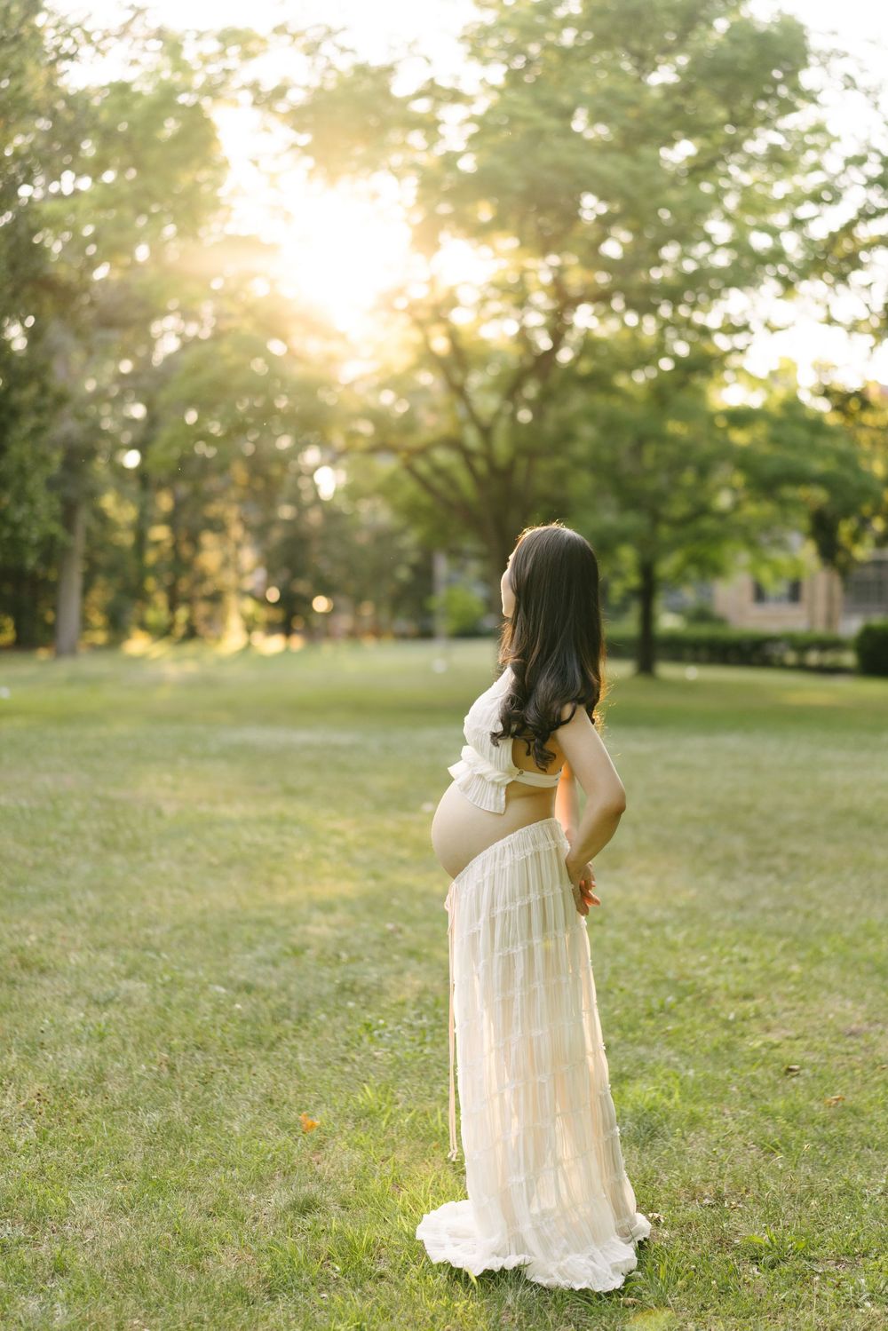 Expectant mother in a lace gown standing in profile in a sunlit park.