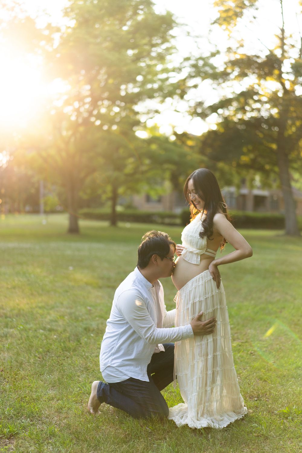 Partner kneeling to hold the expectant mother's belly in a sunlit park.