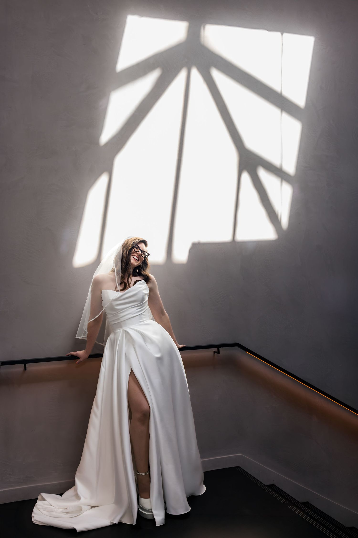 Bride in white strapless gown with slit poses gracefully on stairs beneath geometric window light shadows.