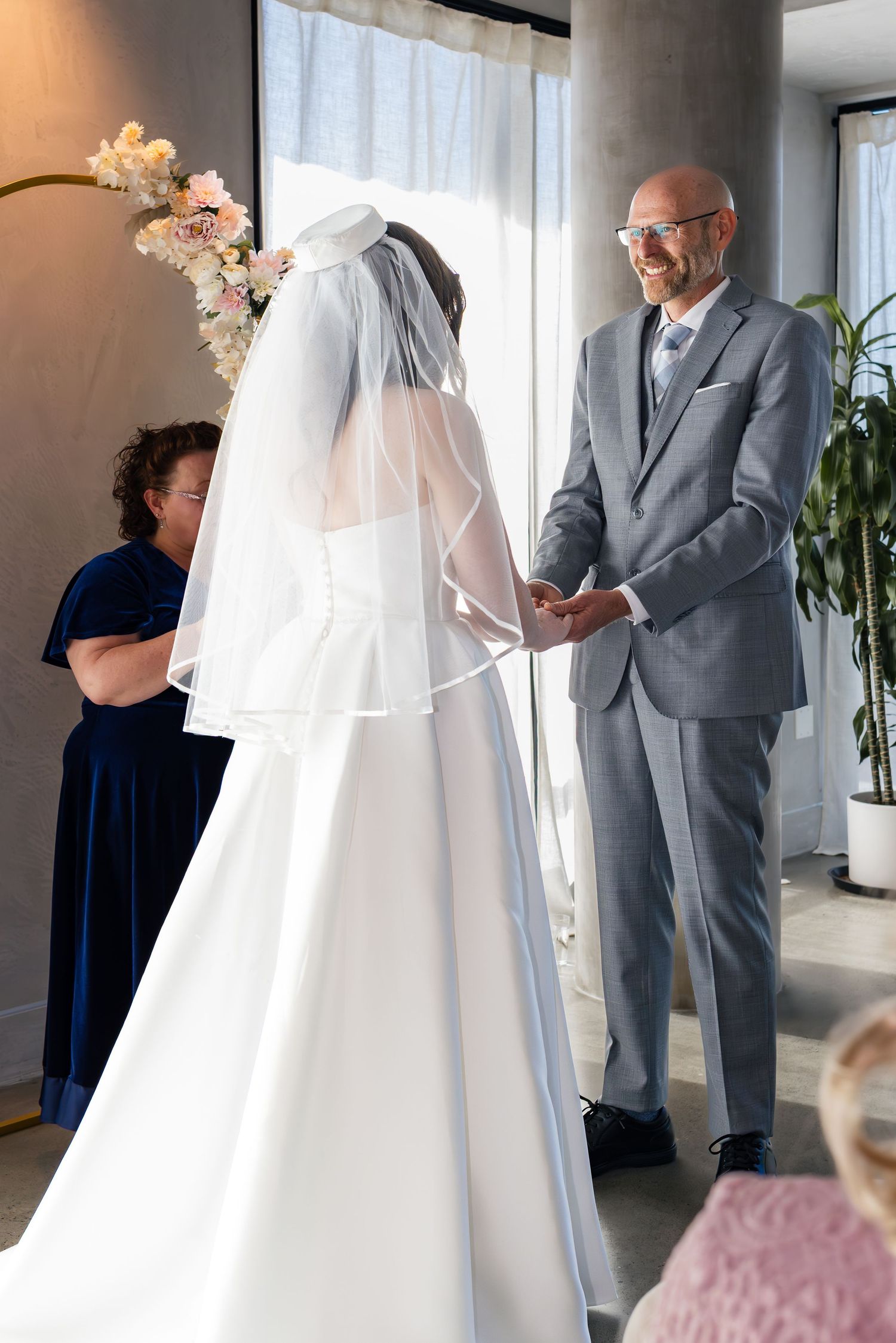 Bride and groom holding hands at indoor wedding ceremony, officiant in blue dress stands behind them.