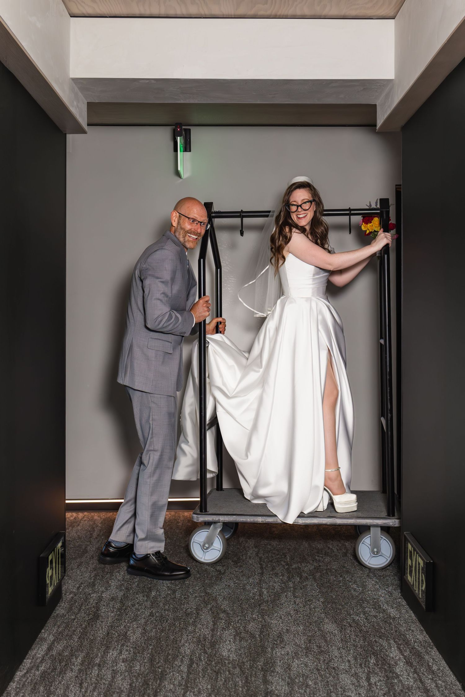 Bride in white gown and groom in gray suit pose playfully on a luggage cart in a hotel corridor.