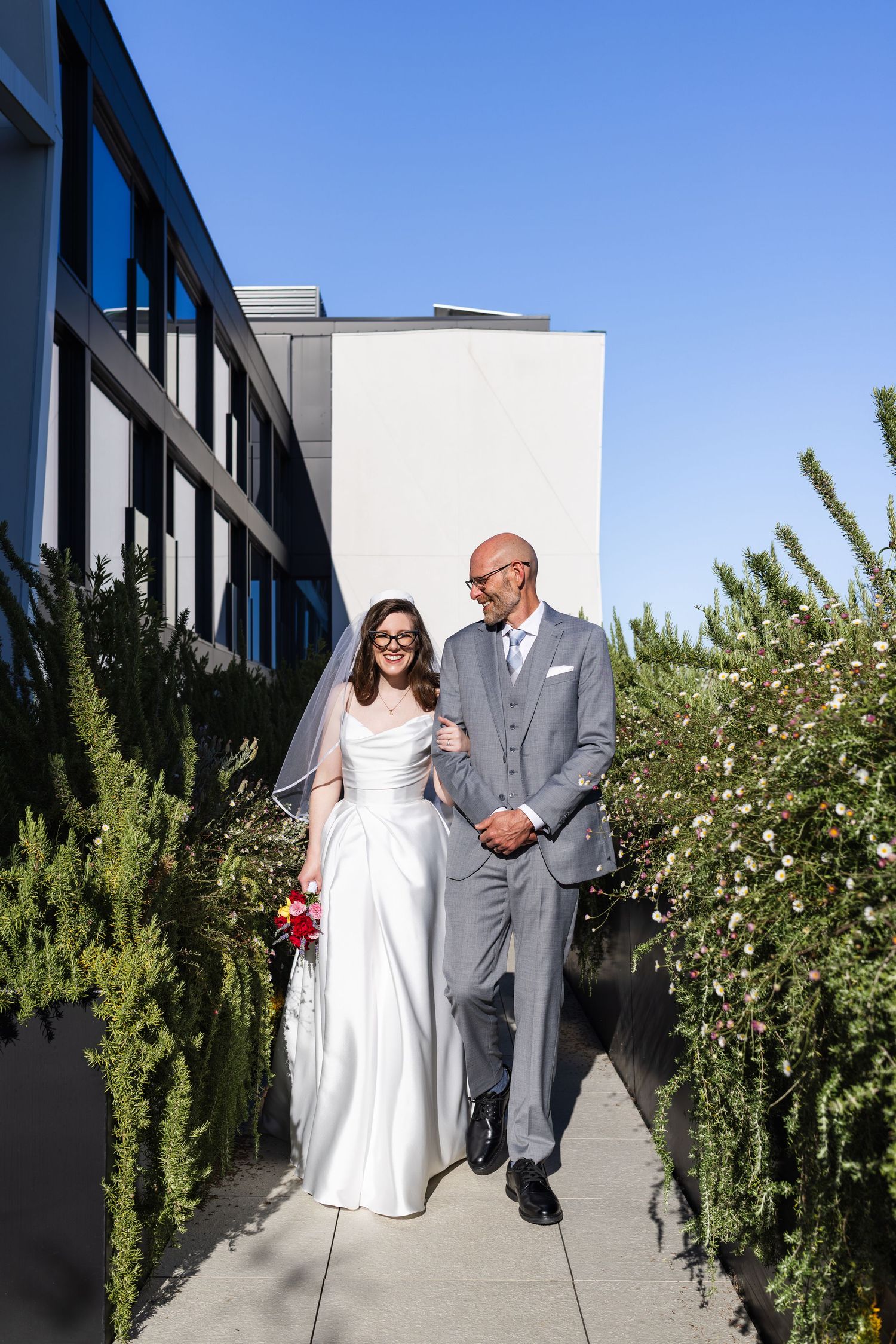 Bride in white gown and veil walks with bald man in gray suit outdoors on sunny day with green shrubs.