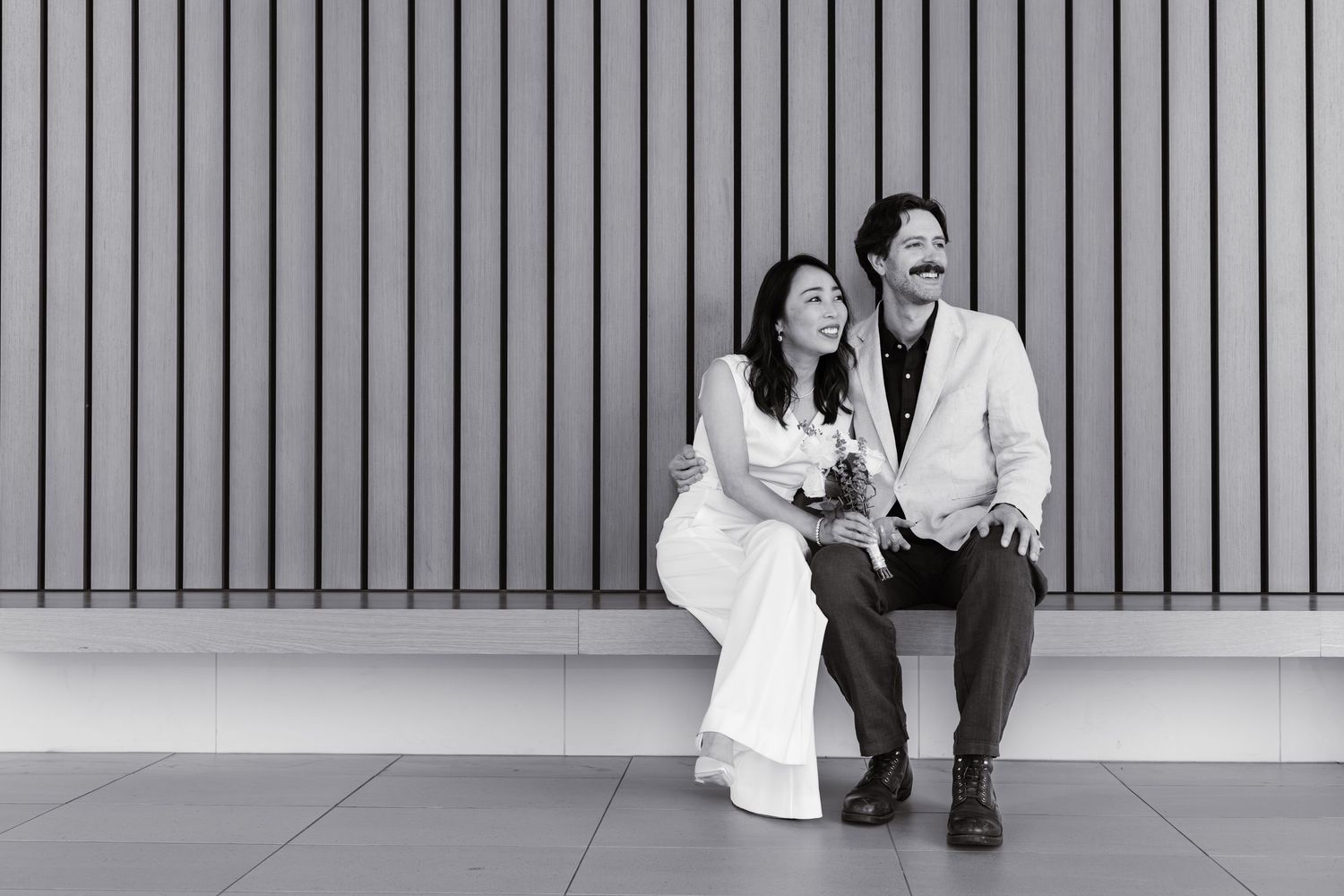 Black and white photo of a couple in wedding attire smiling at each other in a modern building lobby.
