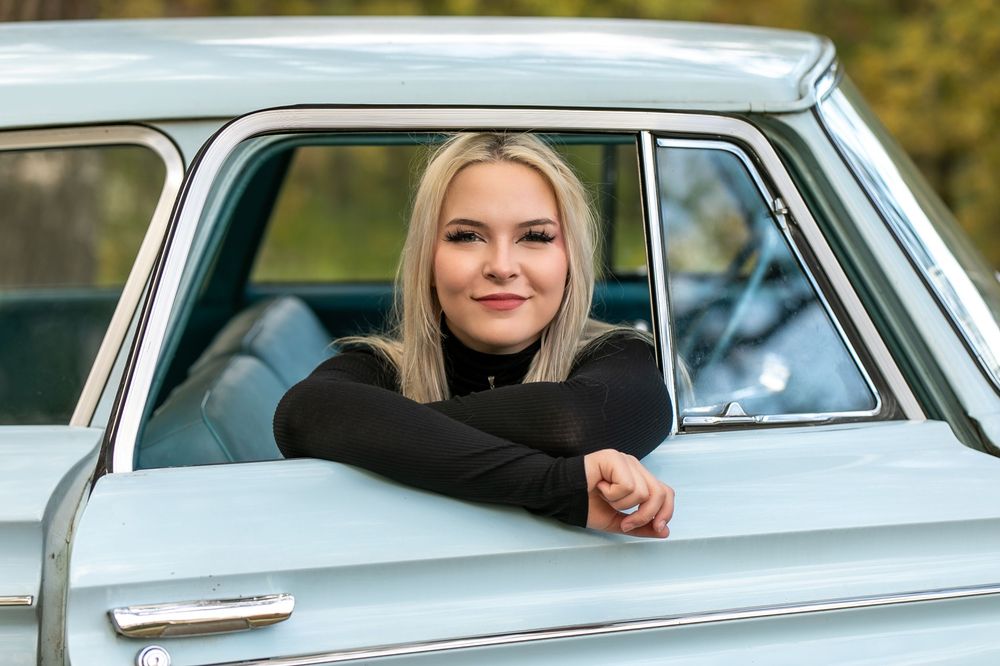Young blonde woman smiling from vintage light blue classic car window, wearing black long-sleeve top outdoors.