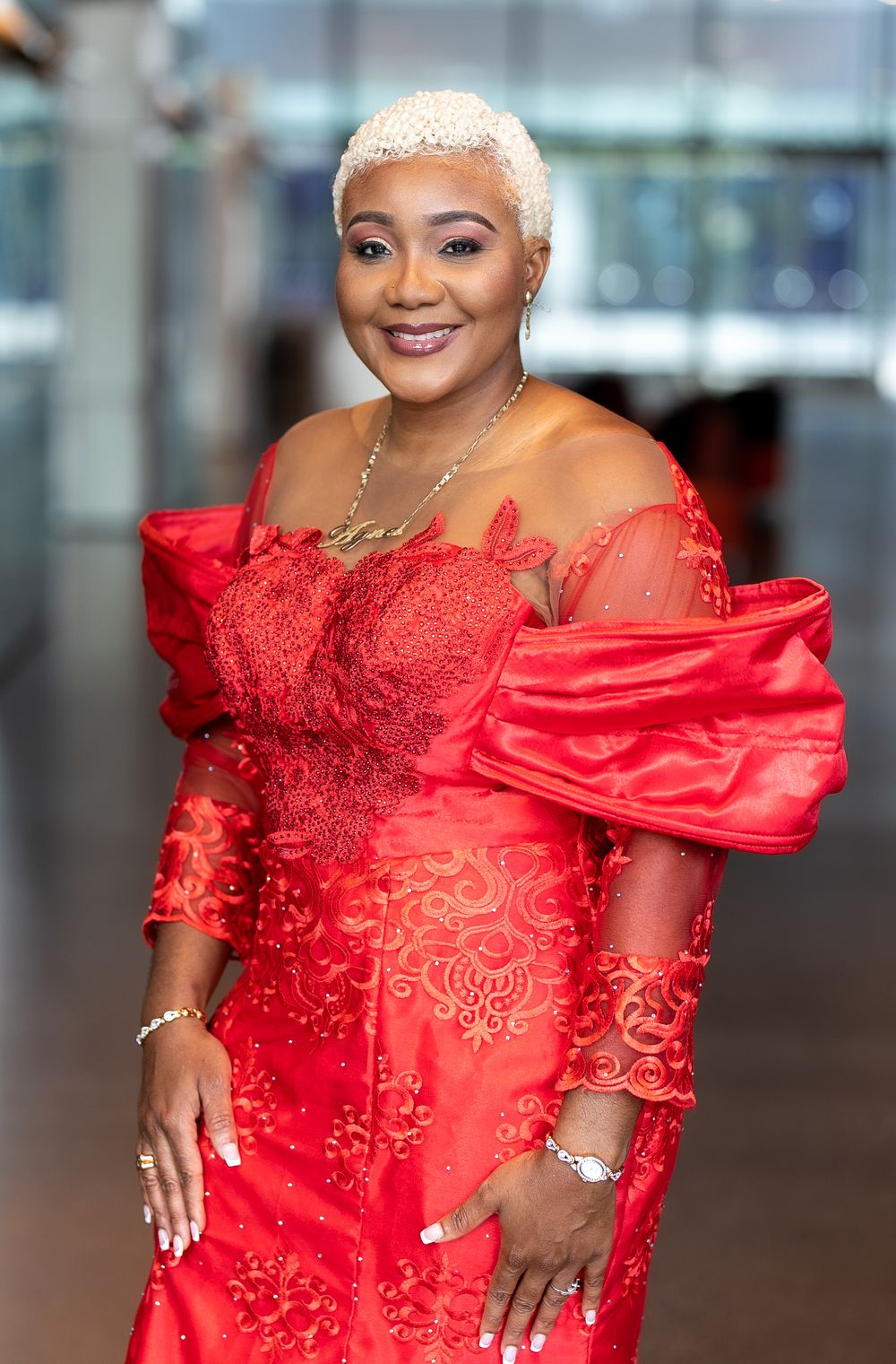 Woman in elegant red lace off-shoulder gown with blonde pixie cut poses gracefully at formal event.