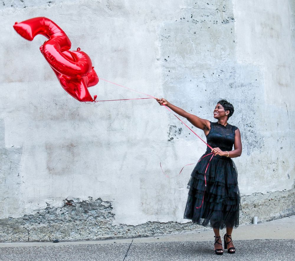 Woman in black dress holding red number balloon against concrete wall outdoors.