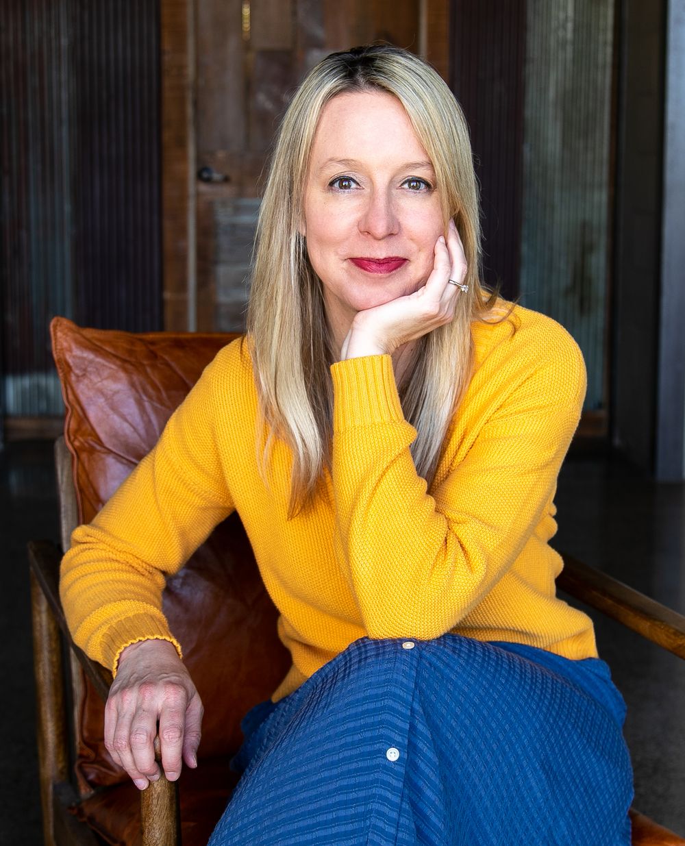 Smiling blonde woman in yellow sweater and blue pants sitting relaxed in brown leather chair.