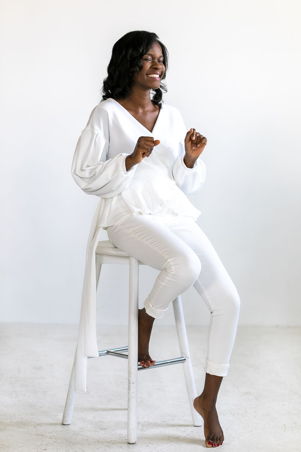 Smiling Black woman in all-white outfit sitting on a white stool against a light background.