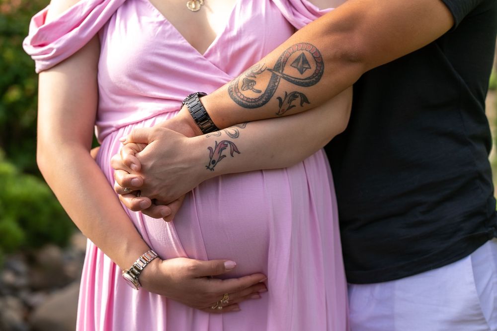 Pregnant woman in pink dress with partner's tattooed arm around her baby bump during maternity photoshoot.