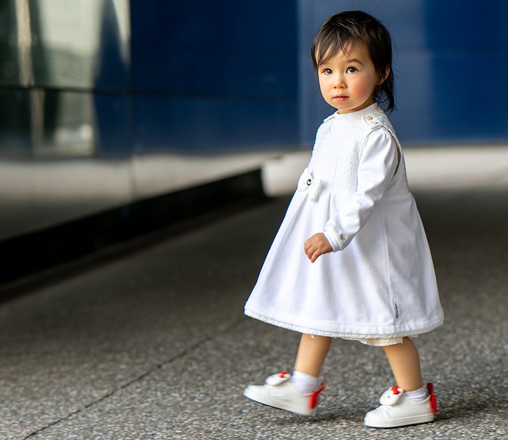 A cute toddler girl in a white dress and red-accented sneakers walks confidently on a sidewalk.