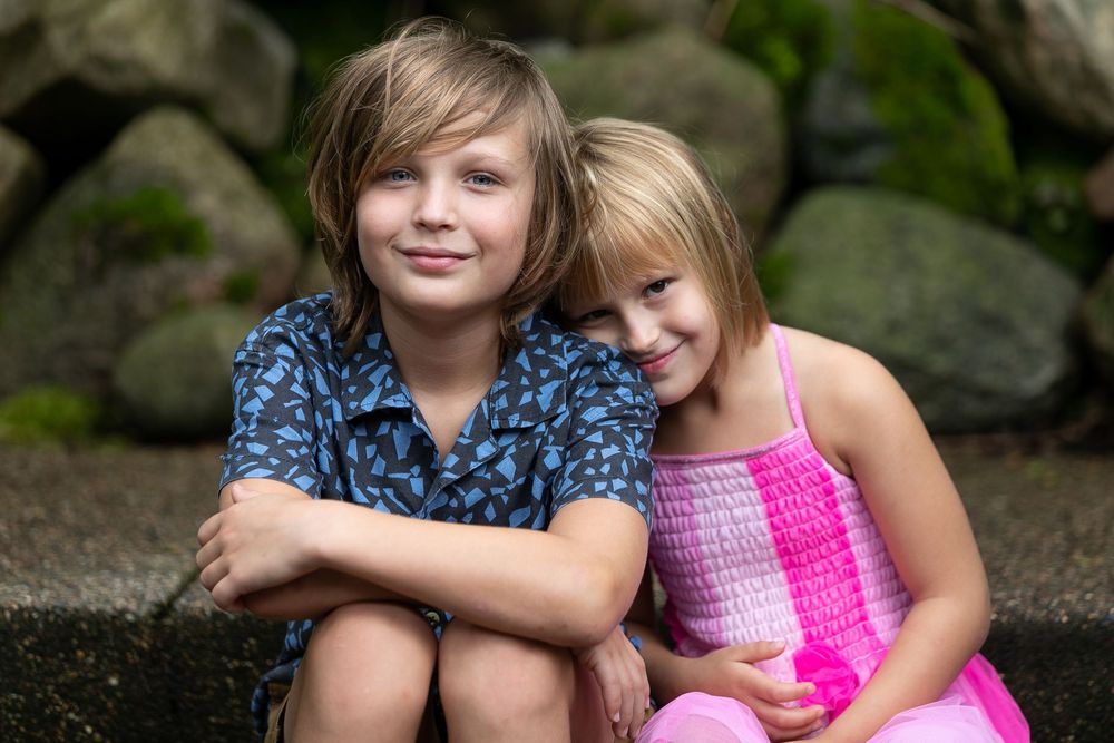 Two smiling children, a boy and girl, sitting together outdoors near stone wall in casual summer clothing.