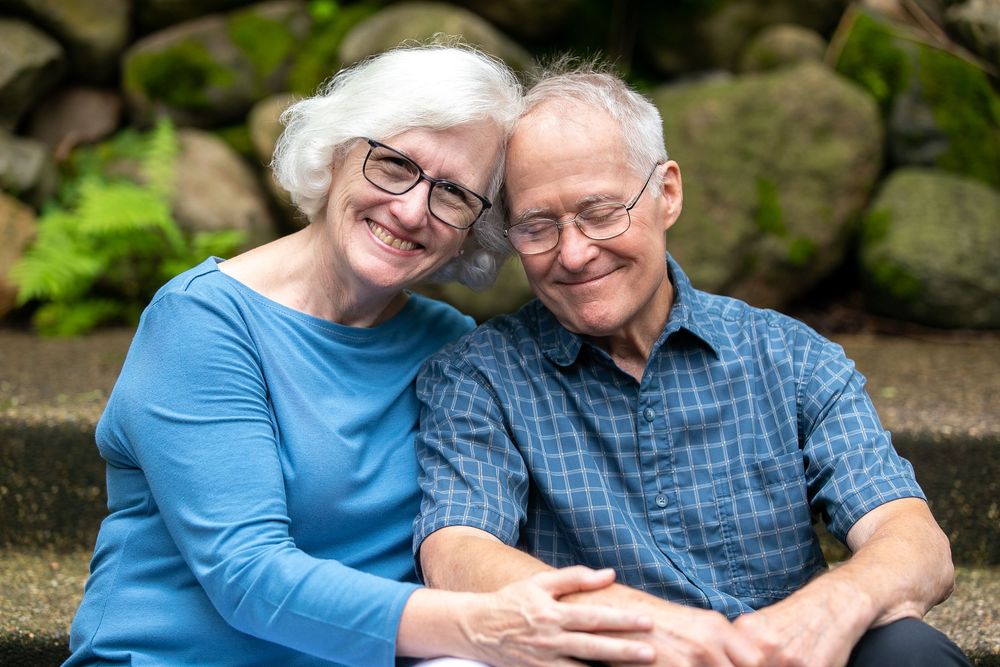 Happy elderly couple smiling and embracing outdoors, sitting together against a stone wall backdrop.