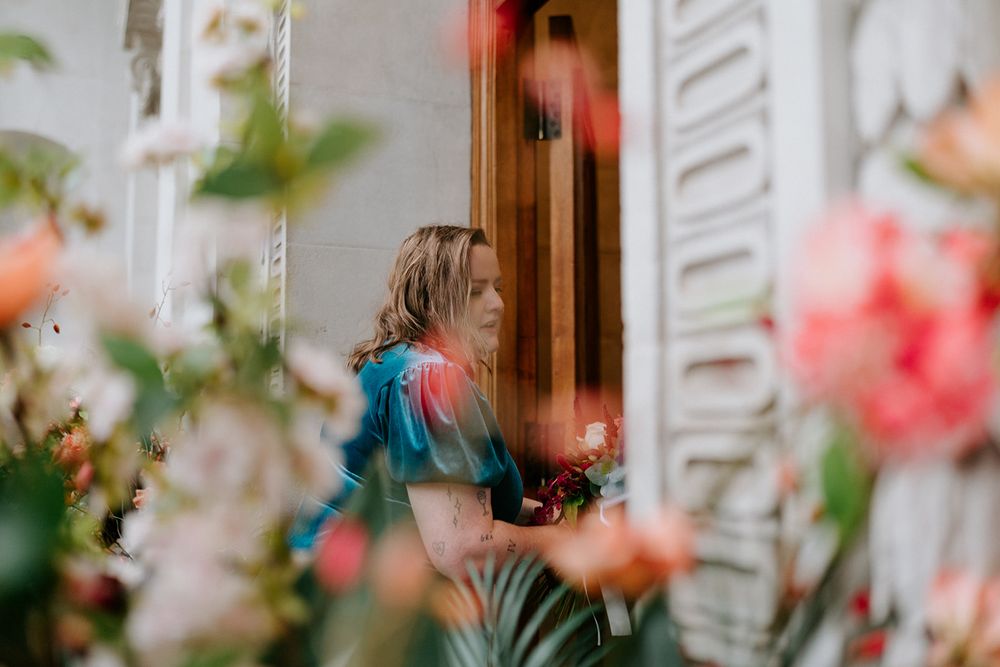 Woman in colorful dress viewed through blooming pink and coral flowers at a floral shop entrance.