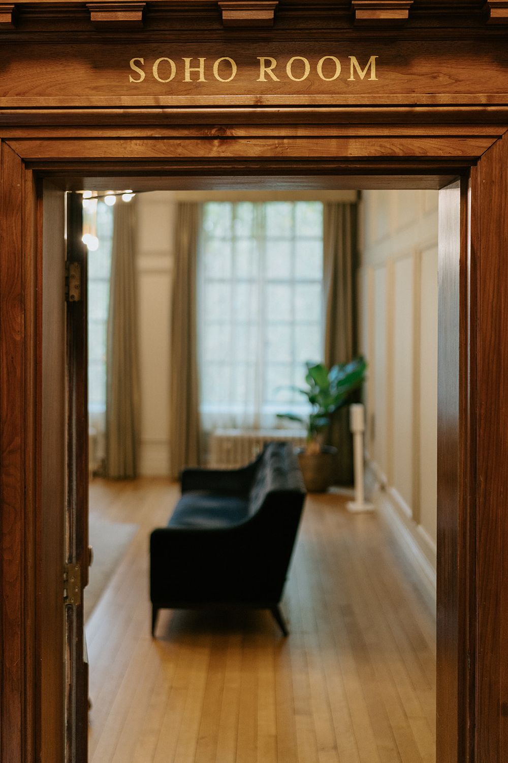 Elegant Soho Room entrance with wooden doorframe, dark velvet sofa, hardwood floors, and large windows with draping curtains.