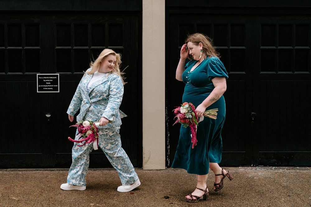 Two women in colorful outfits holding bouquets laugh together outside black doors.