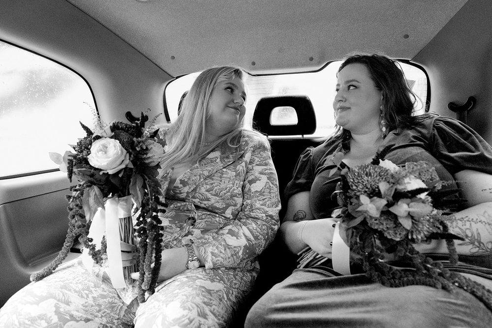 Two brides in wedding attire smile at each other while holding bouquets in the back of a car, black and white photo.
