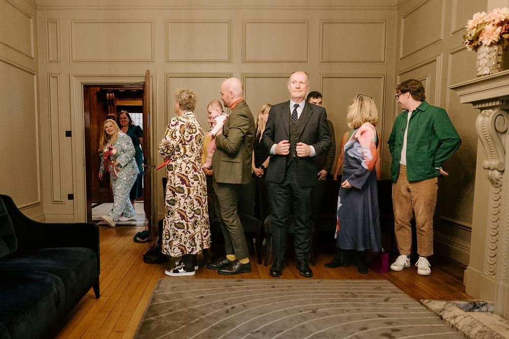 Elegantly dressed wedding guests mingle and chat in a classic wood-paneled reception room.