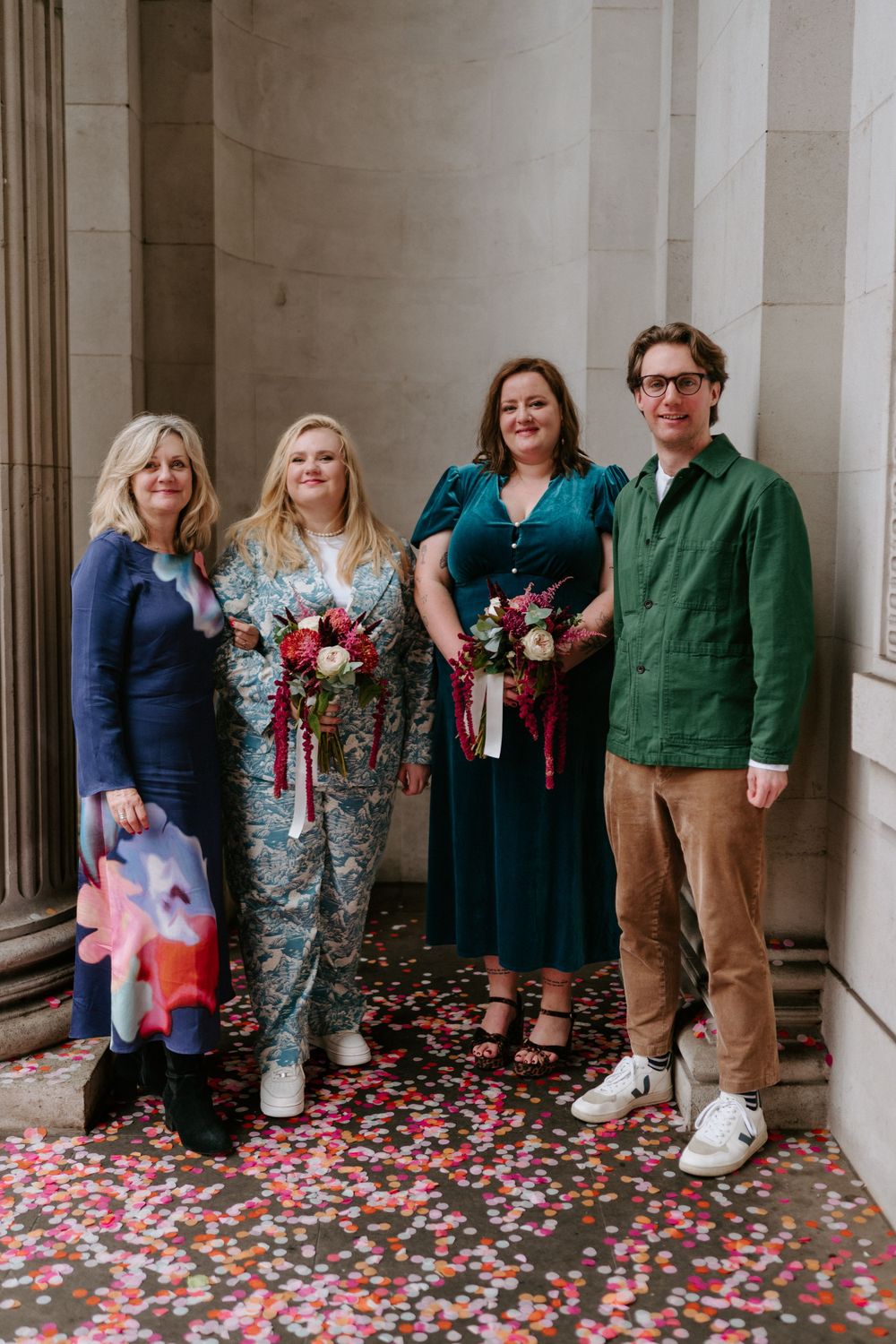 Four wedding guests pose together on confetti-covered steps, two holding colorful floral bouquets.
