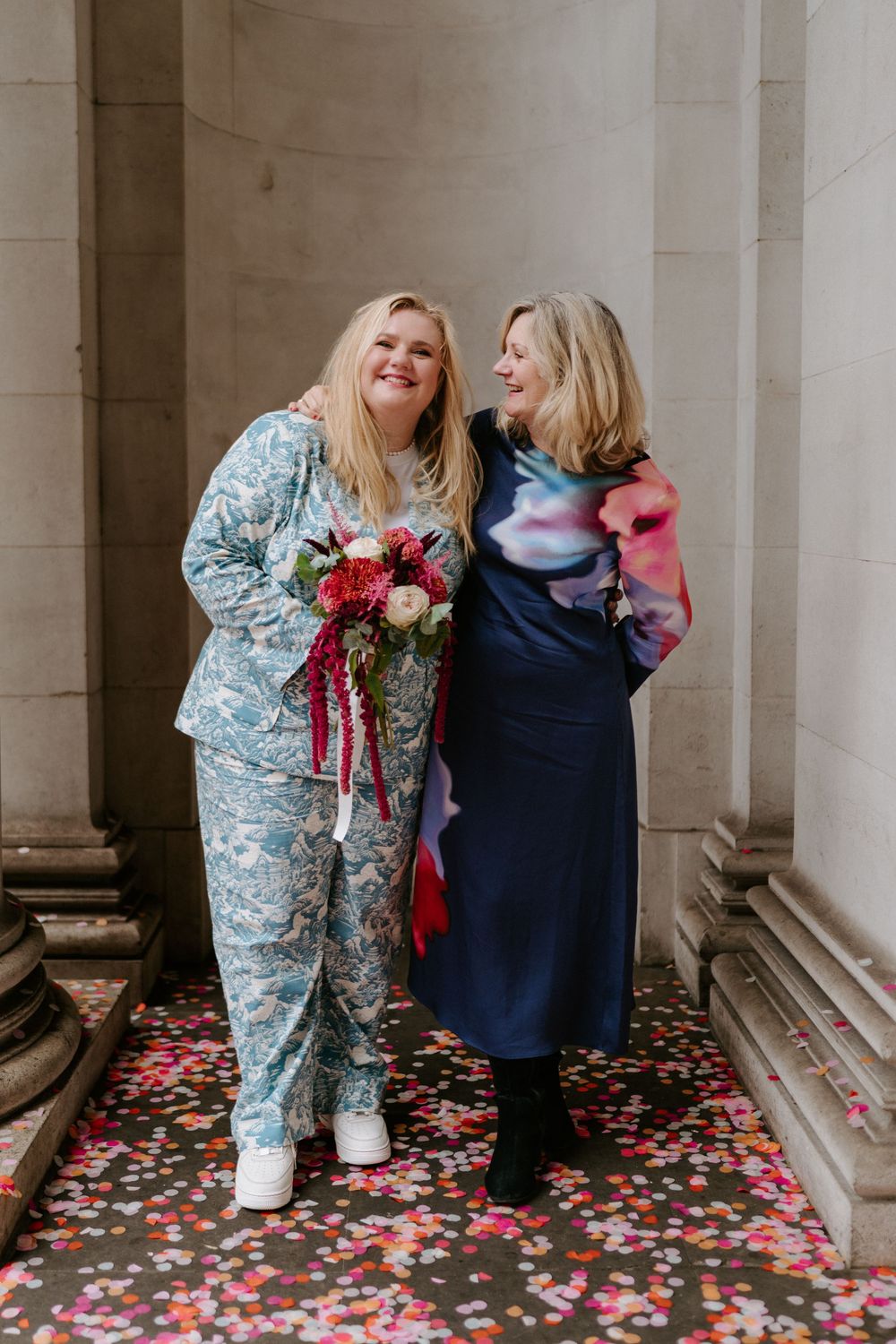 Two women smile together among colorful confetti at a wedding, one holding a red and pink bouquet.