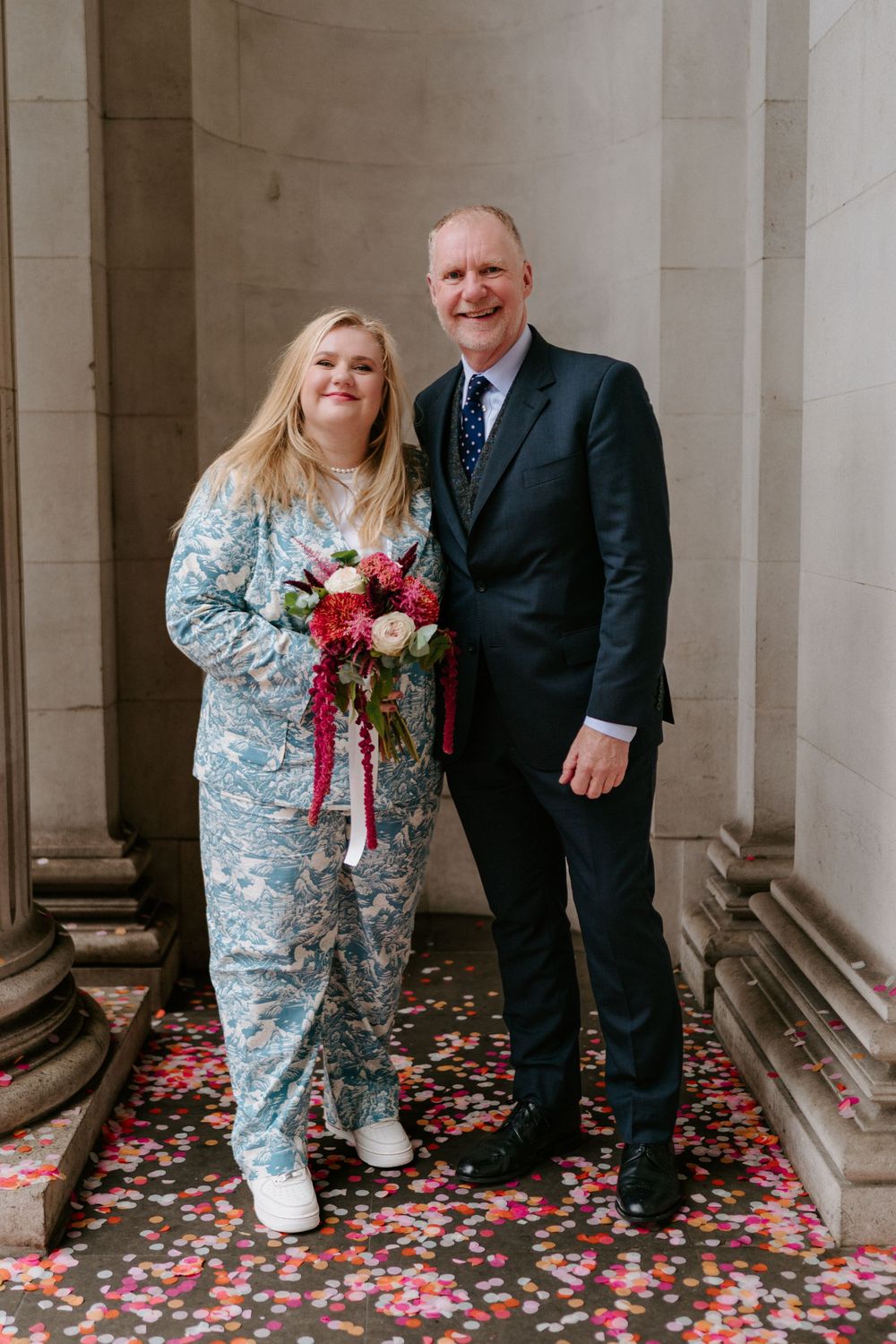 A bride in a floral pantsuit holds a bouquet beside a man in a navy suit on a confetti-covered floor.