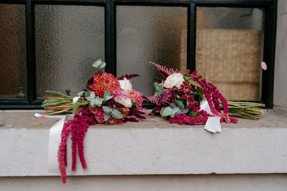 Two elegant bridal bouquets with deep red amaranthus, pink dahlias, and white roses rest on a ledge.