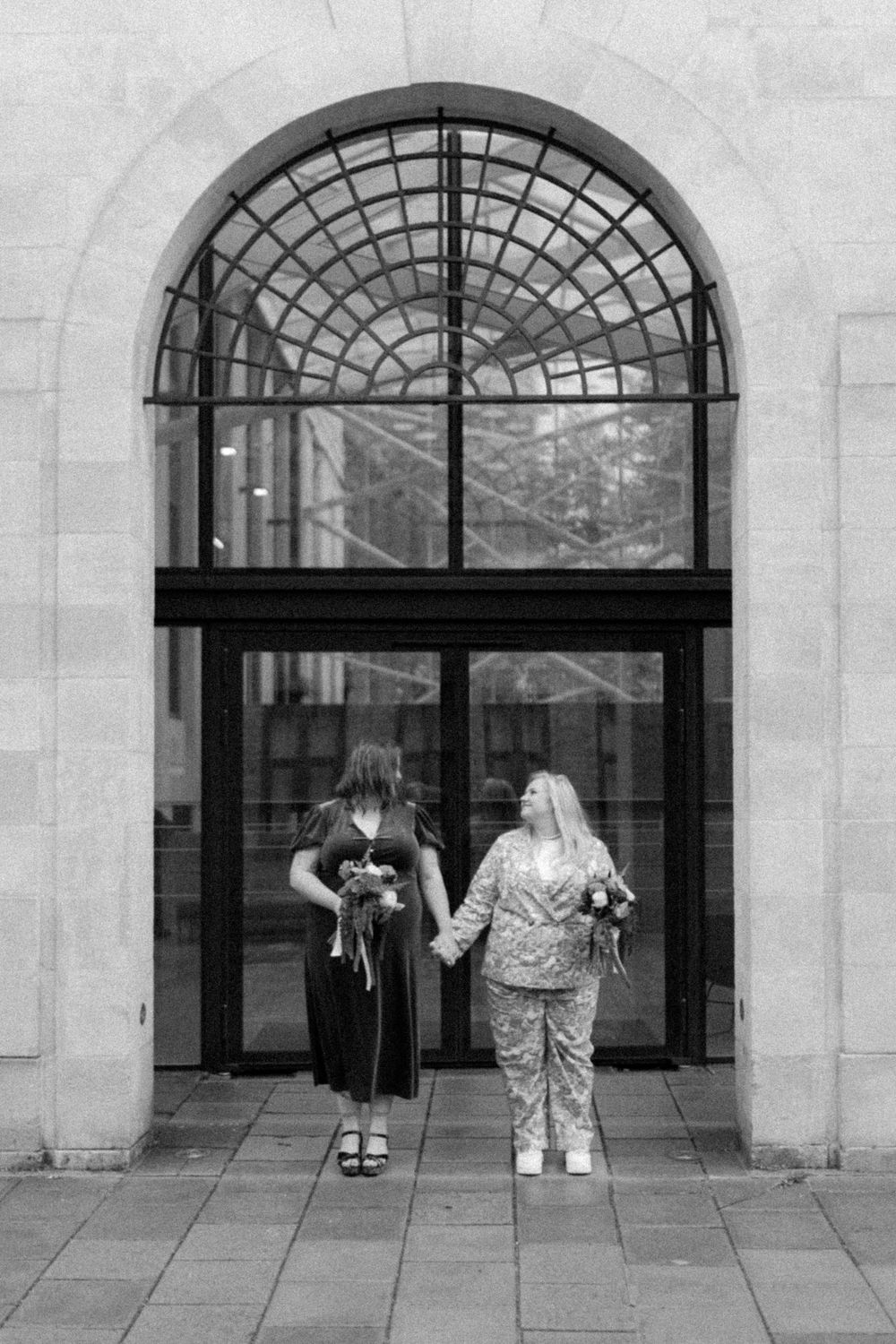 Two women hold hands facing arched glass door in black and white wedding photo, each carrying small bouquets.