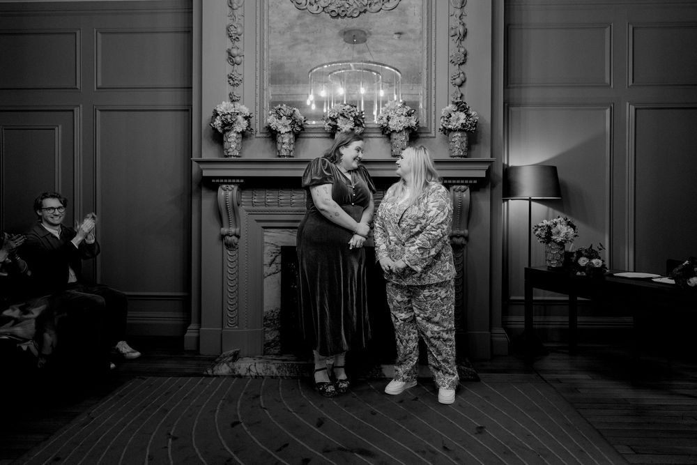 Black and white photo of two women holding hands at an elegant indoor wedding ceremony by a fireplace.