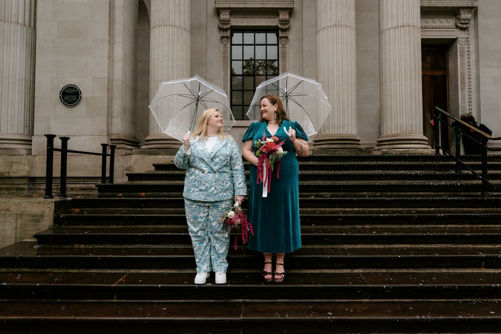 Two brides holding umbrellas and bouquets on stone steps outside a grand building on their wedding day.