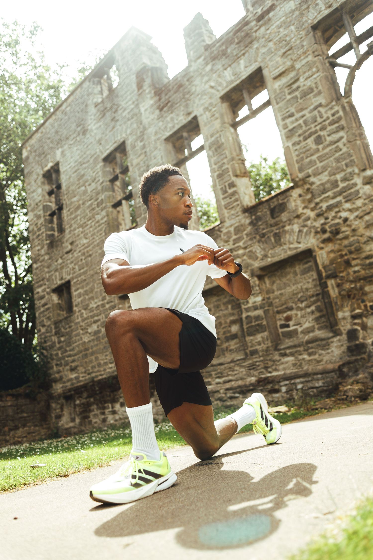 Athletic man in white t-shirt and black shorts doing lunges outdoors near historic stone ruins.