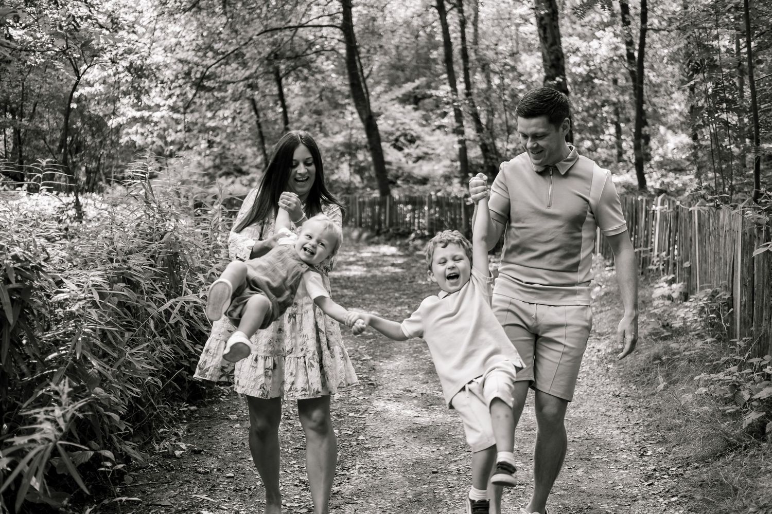 Black and white photo of a happy family walking on a forest path, parents swinging child between them.