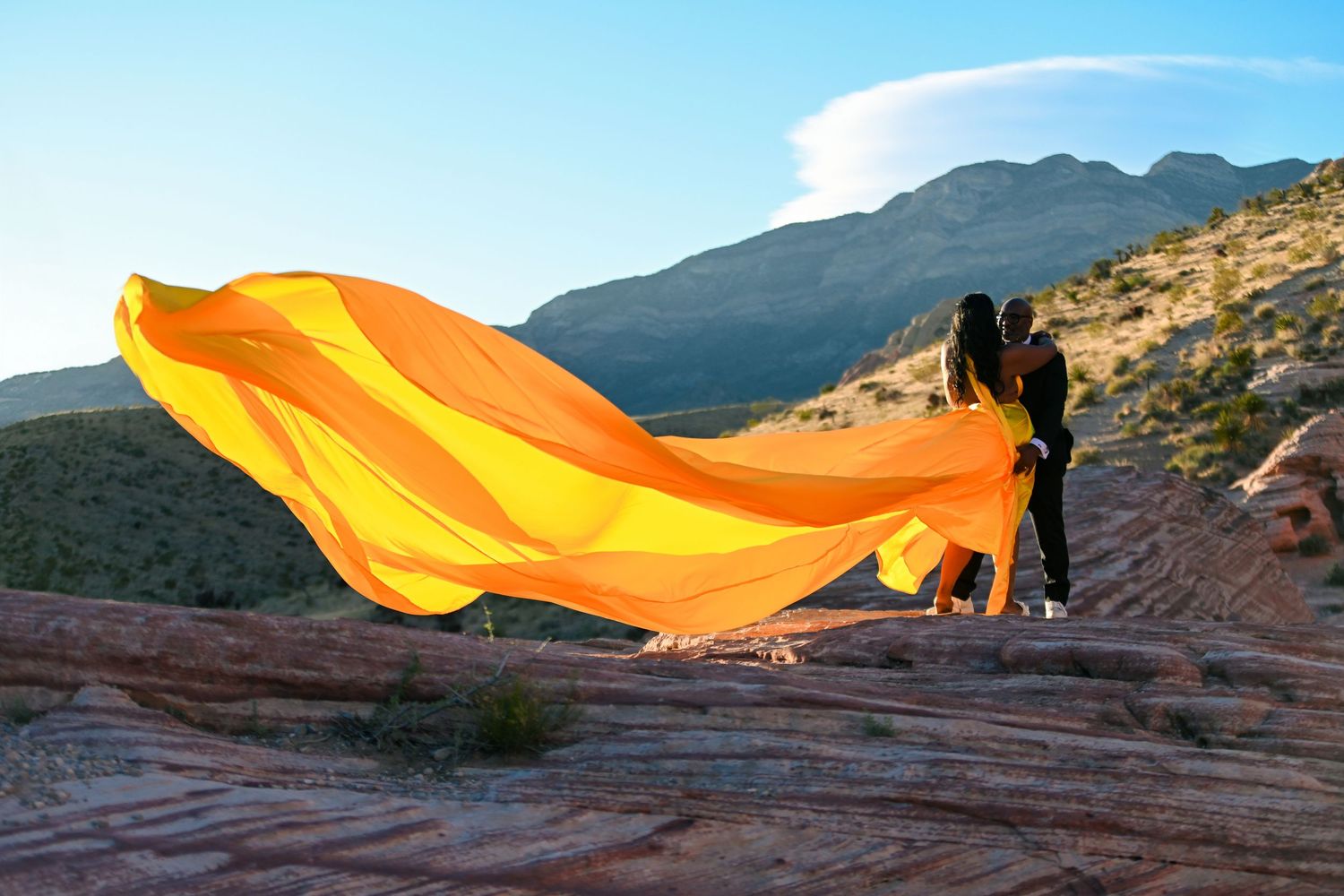 Couple embracing in desert landscape with flowing orange fabric billowing in wind against mountain backdrop.