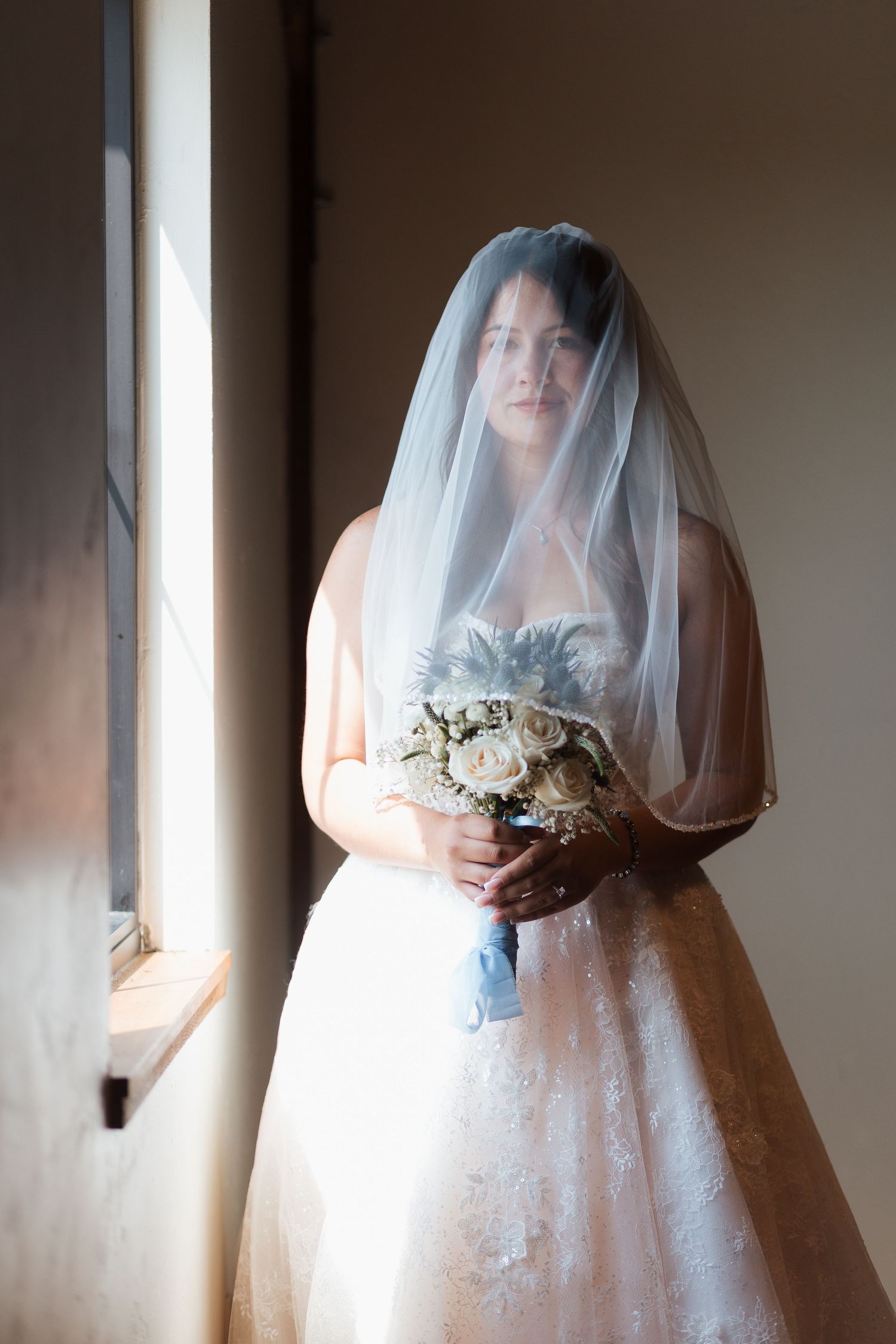 Bride in lace wedding dress and veil holding white rose bouquet standing by window in soft natural light.