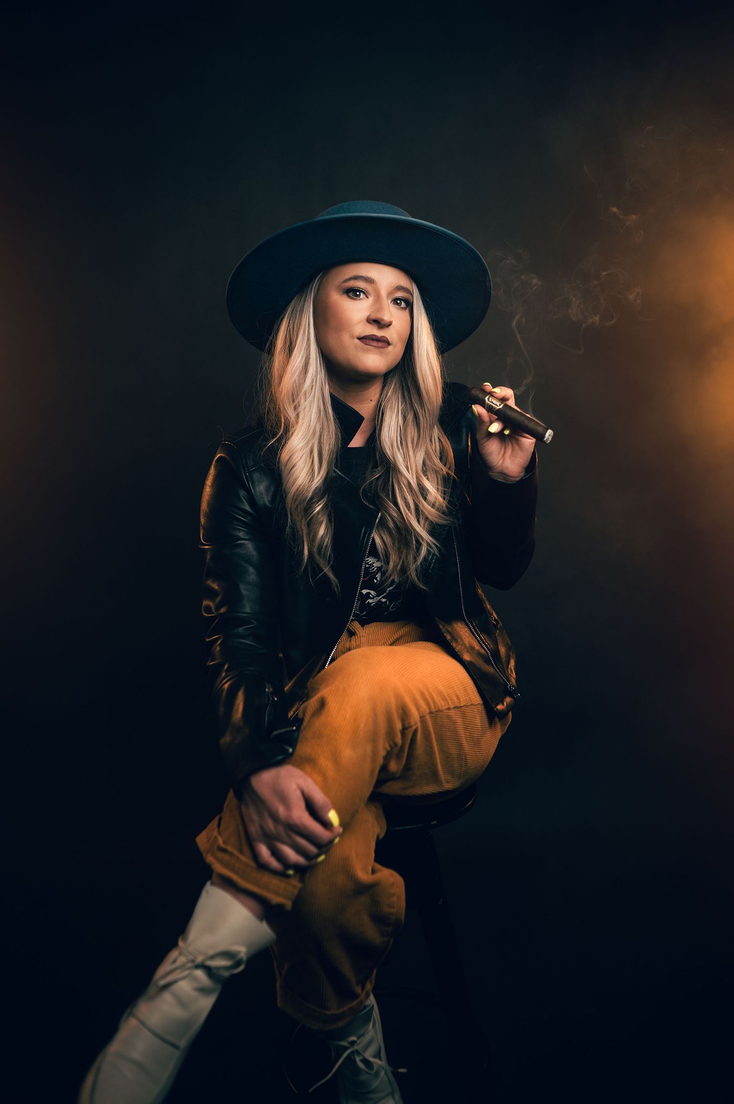 Blonde woman in wide-brim hat, leather jacket, and tan pants poses confidently while smoking a cigar in dramatic lighting.