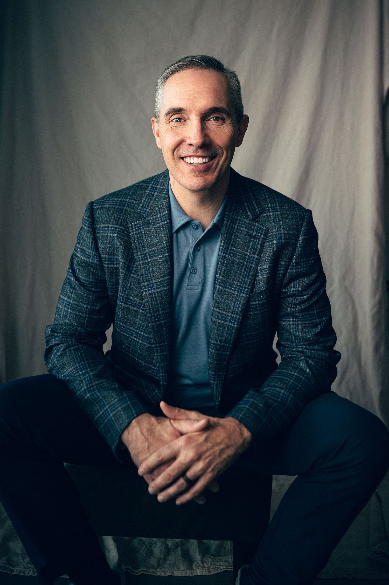Smiling middle-aged man in plaid blazer and blue shirt seated confidently against gray backdrop.