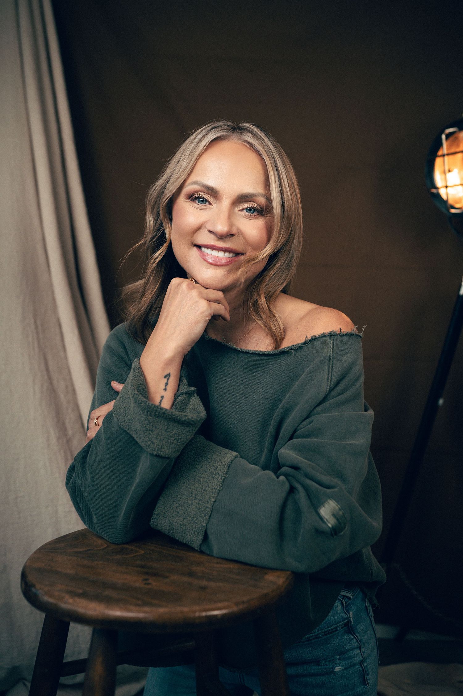 Smiling woman with blonde hair poses on a wooden stool in a green off-shoulder sweater and jeans in a studio.