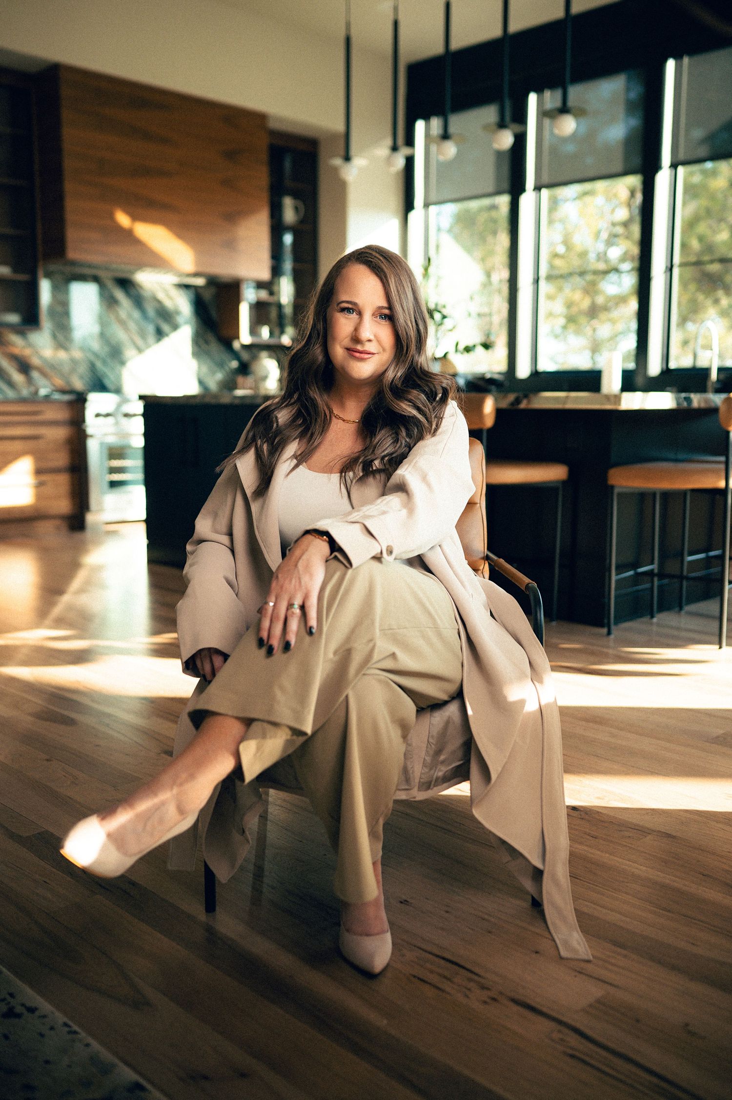 Woman in beige trench coat and wide-leg pants seated confidently in a modern luxury kitchen interior.