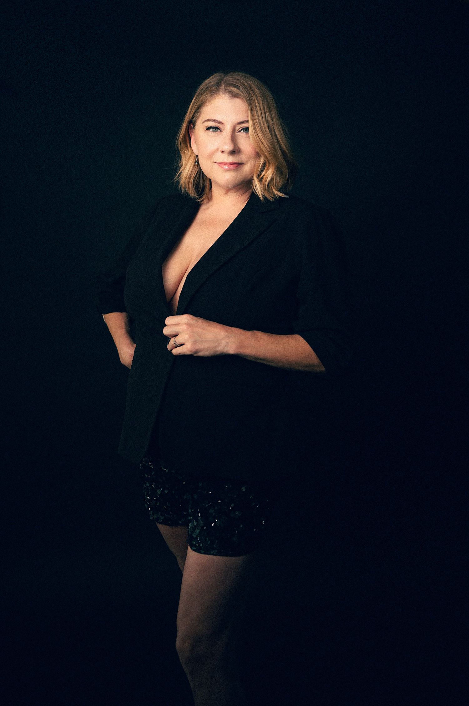 Woman in black blazer and sequin shorts posing confidently against dark studio background.