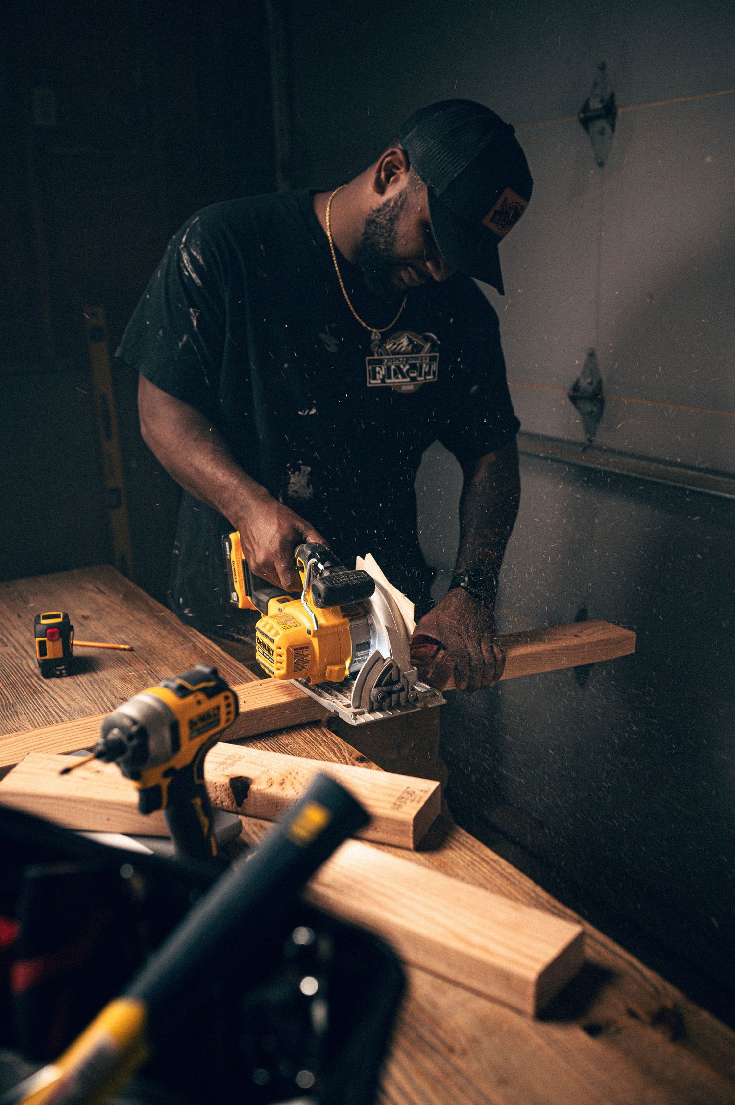 A man uses a DeWalt circular saw on wood at a workshop bench surrounded by power tools in a dimly lit workspace.