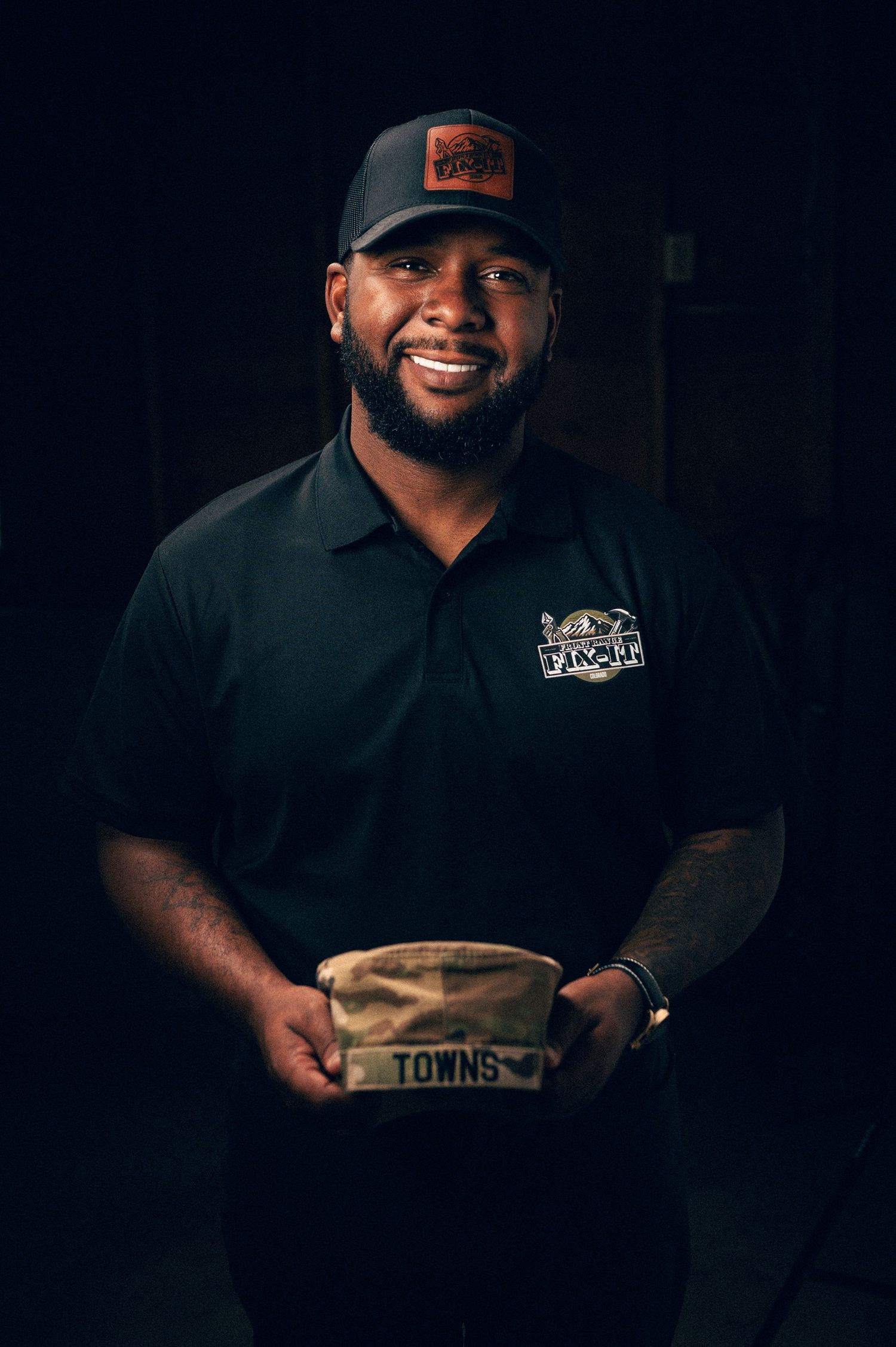 Smiling man in black polo shirt and cap holding a branded Towns bag against dark background.